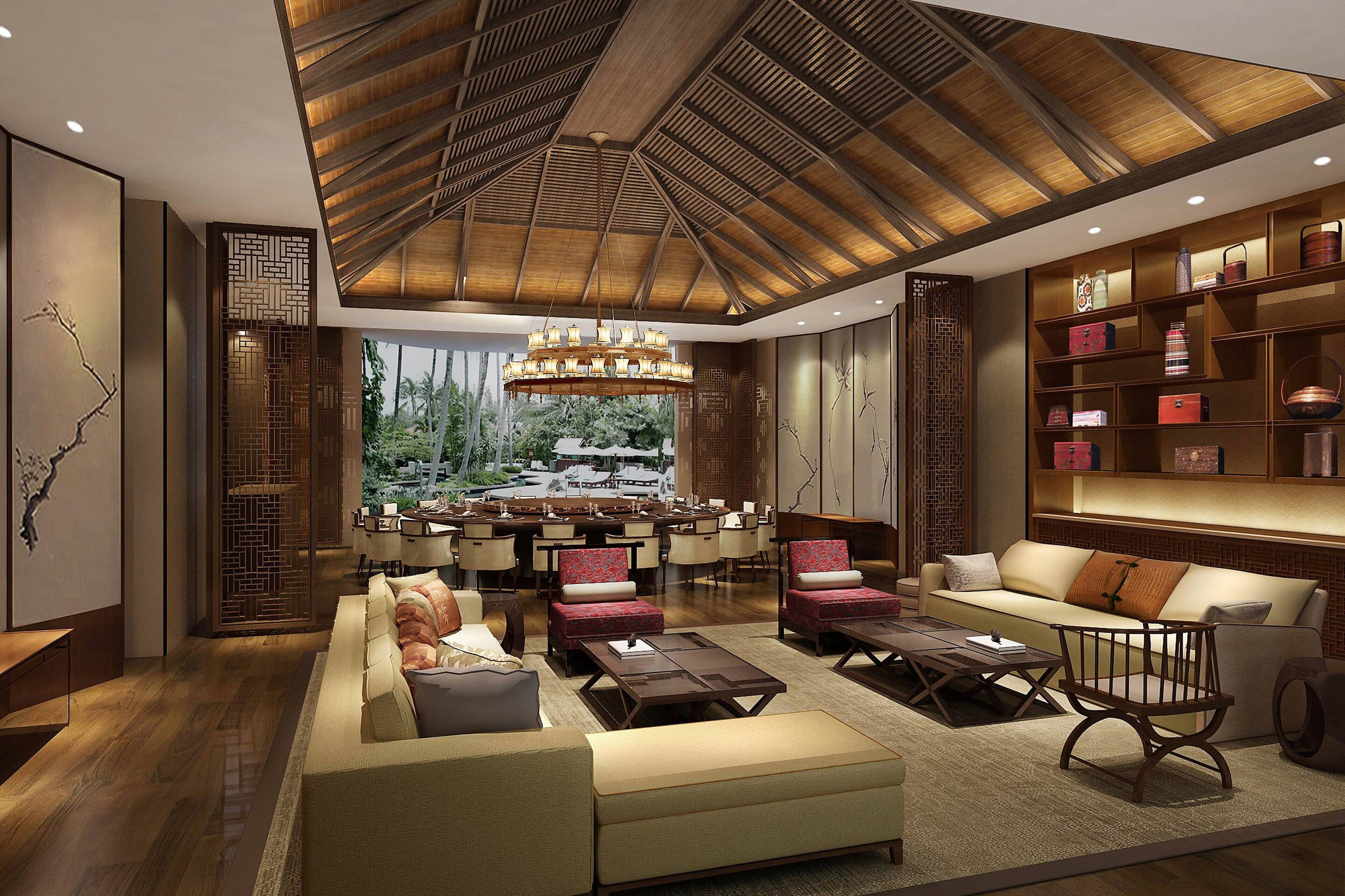 A spacious living area with a high ceiling, wooden beams, and a central chandelier. There are multiple seating options including sofas and chairs, and a bookshelf filled with decorative items.
