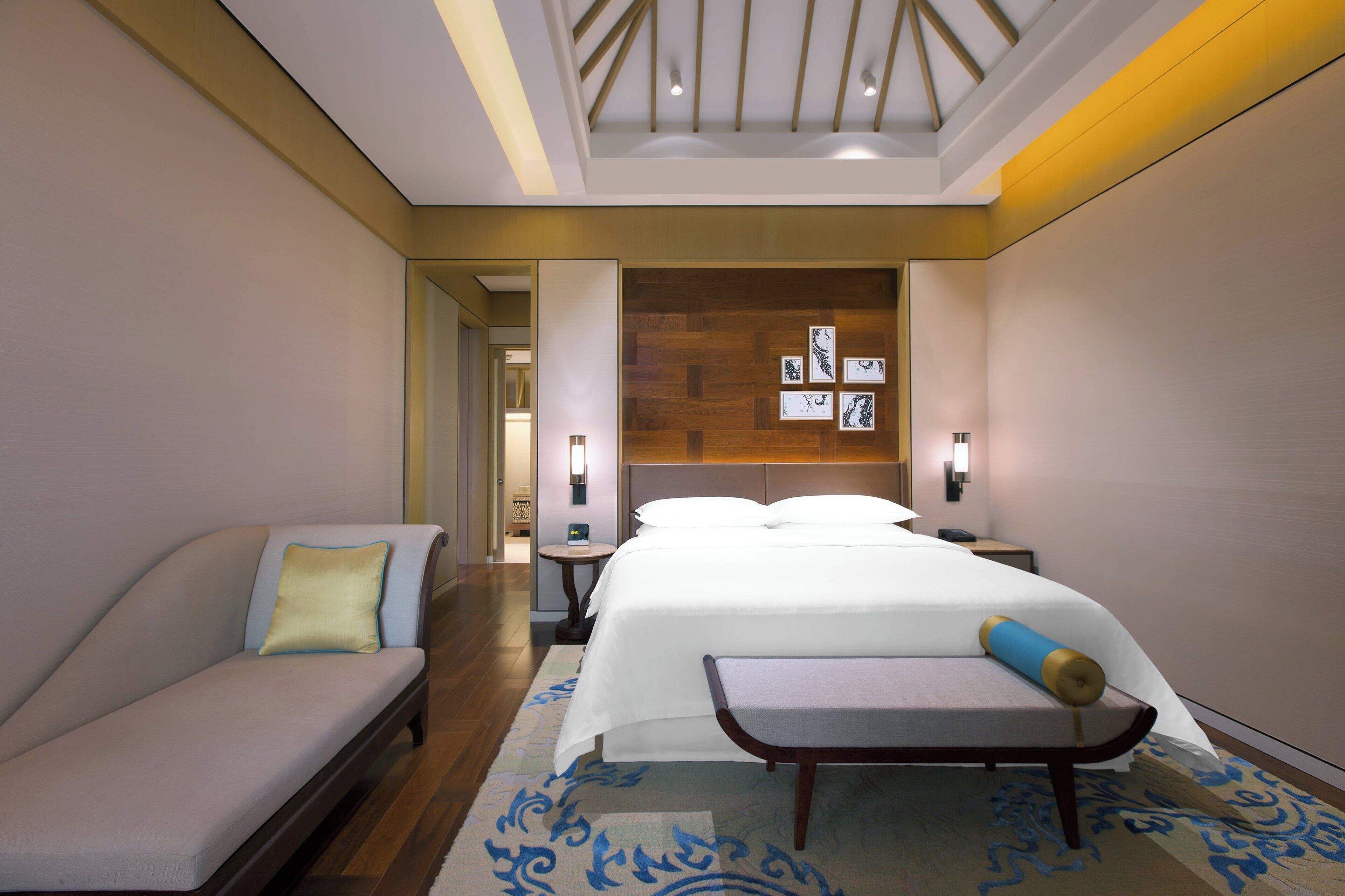 A hotel room with a bed, a sofa, a bench, a wooden headboard, and a ceiling with a skylight.