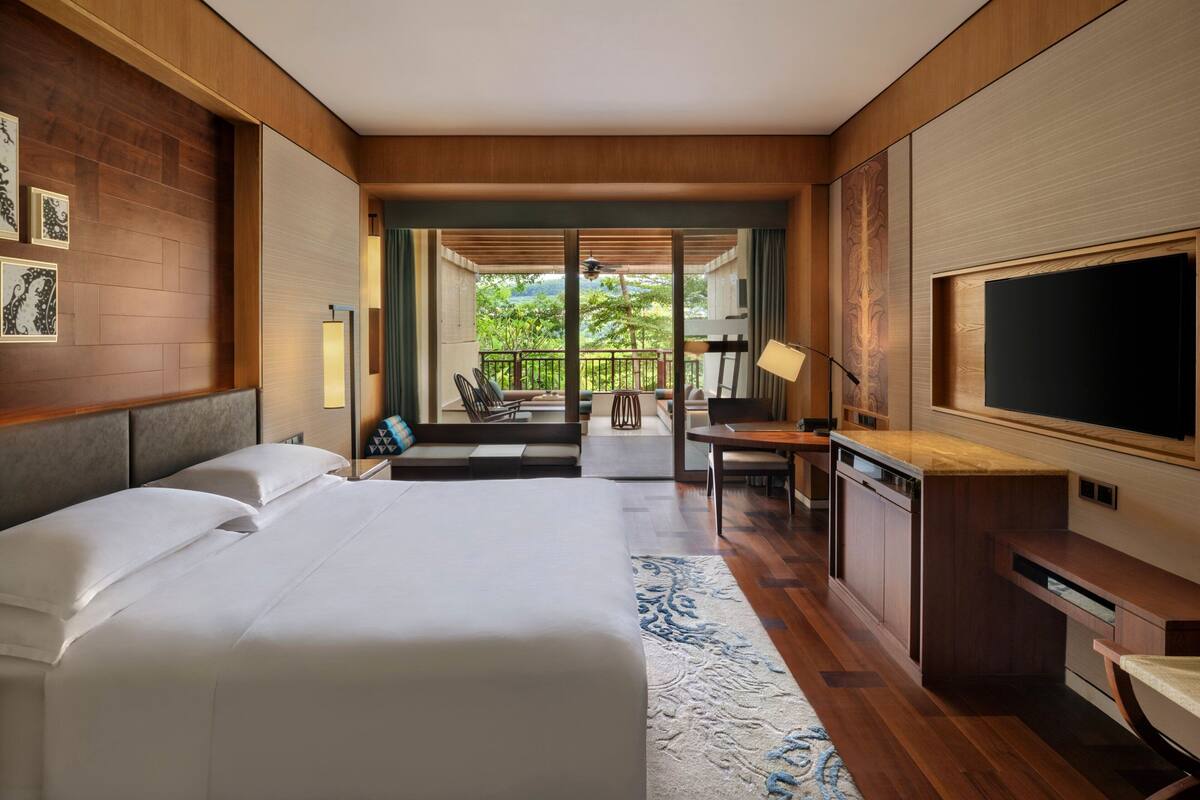 A hotel room with a large bed, a desk with a TV, and a balcony with seating.