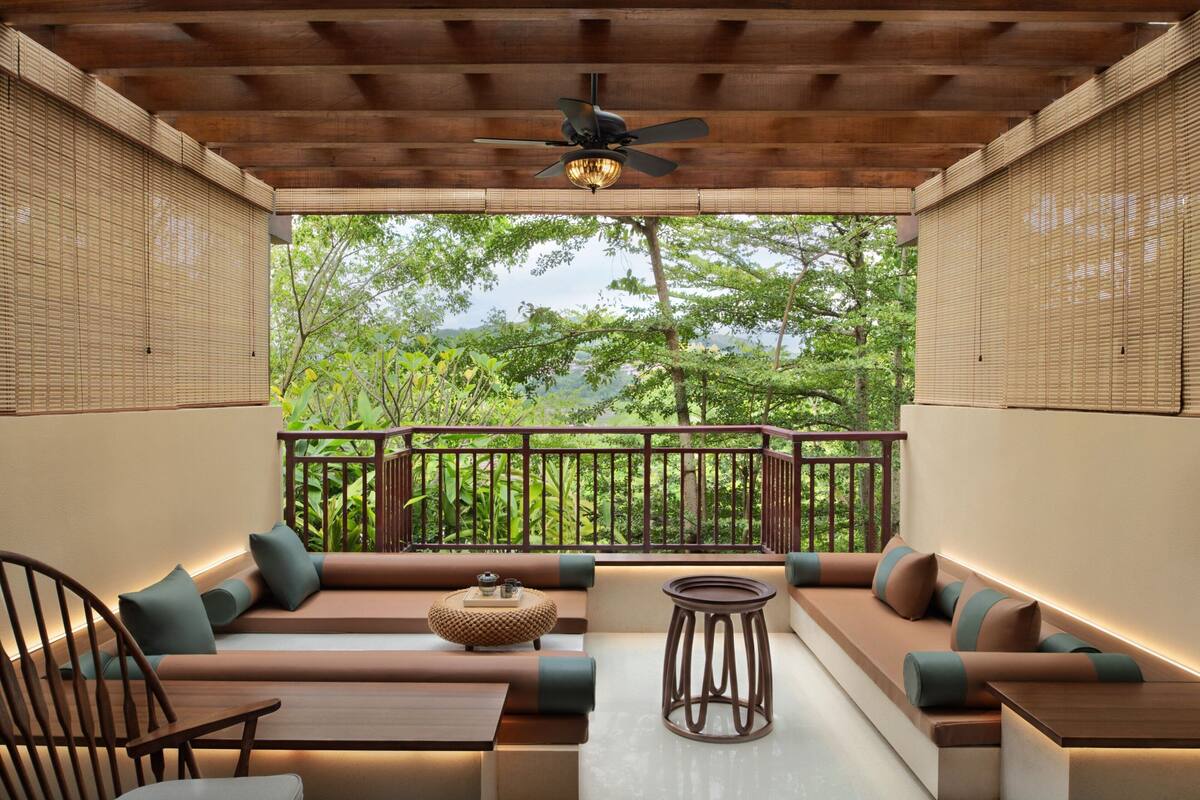 A modern outdoor seating area with a ceiling fan, wooden furniture, and a view of lush greenery.