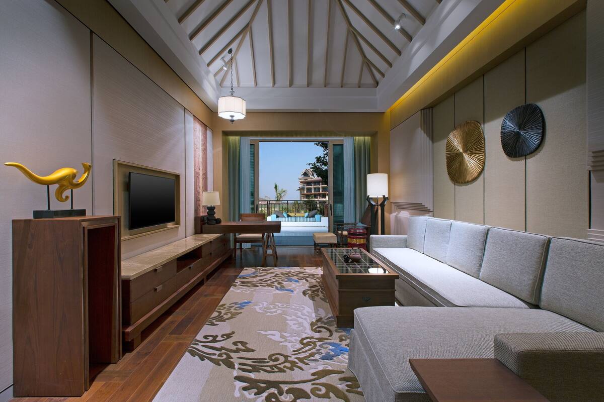 A modern hotel room with a large flat-screen TV, a sofa, a dining area, and a view of a building through a large window.