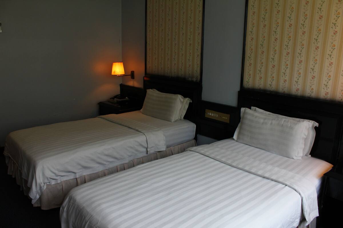 A hotel room with two single beds, a nightstand with a lamp, and floral-patterned curtains.