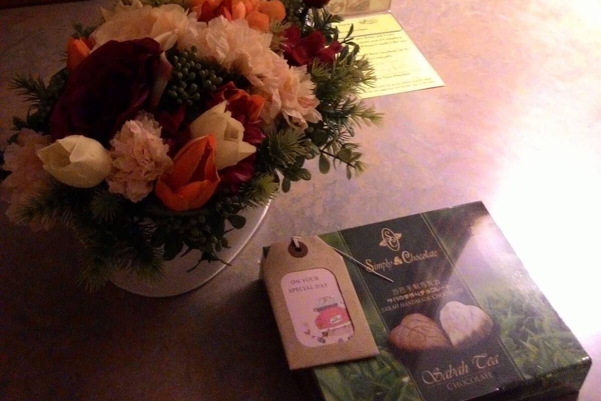 A hotel room with a neatly made bed, a floral arrangement on the side, and a chocolate gift box.