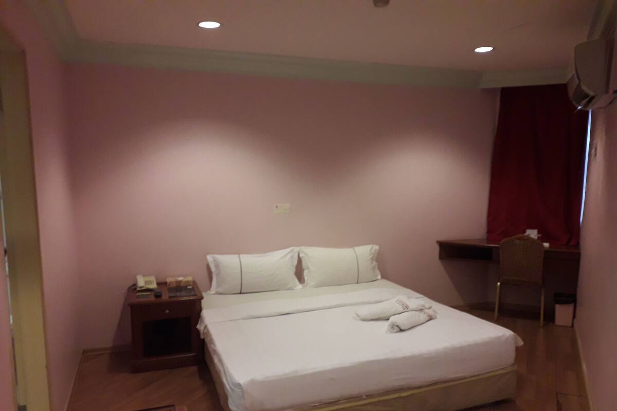 A neatly made bed with white linens, a wooden nightstand with a phone, a desk with a chair, and a red curtain.
