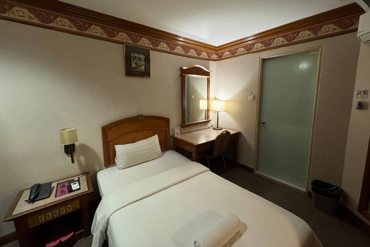 A hotel room with a bed, a desk, a chair, a lamp, a telephone, and a door.