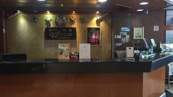 Un mostrador de recepción de hotel con una pantalla digital, un cartel que dice 'Feliz Navidad' y varios avisos y tarjetas en la pared.