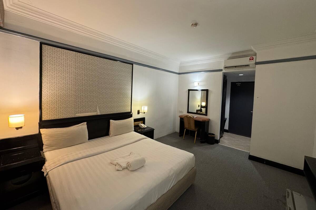 A hotel room with a large bed, a desk with a chair, a mirror, and a small table with a lamp.