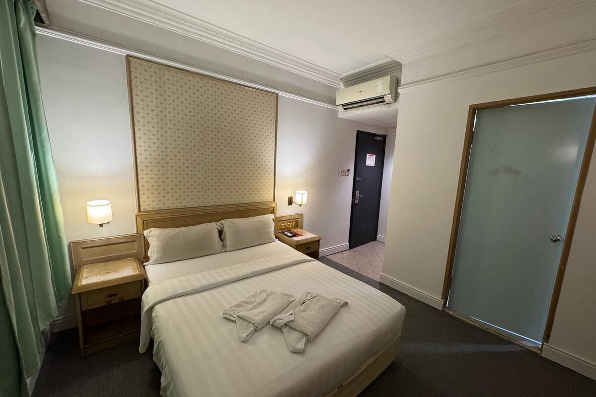 A hotel room with a large bed, two bedside tables, a nightstand, an air conditioning unit, a door, and a wall with a patterned wallpaper.