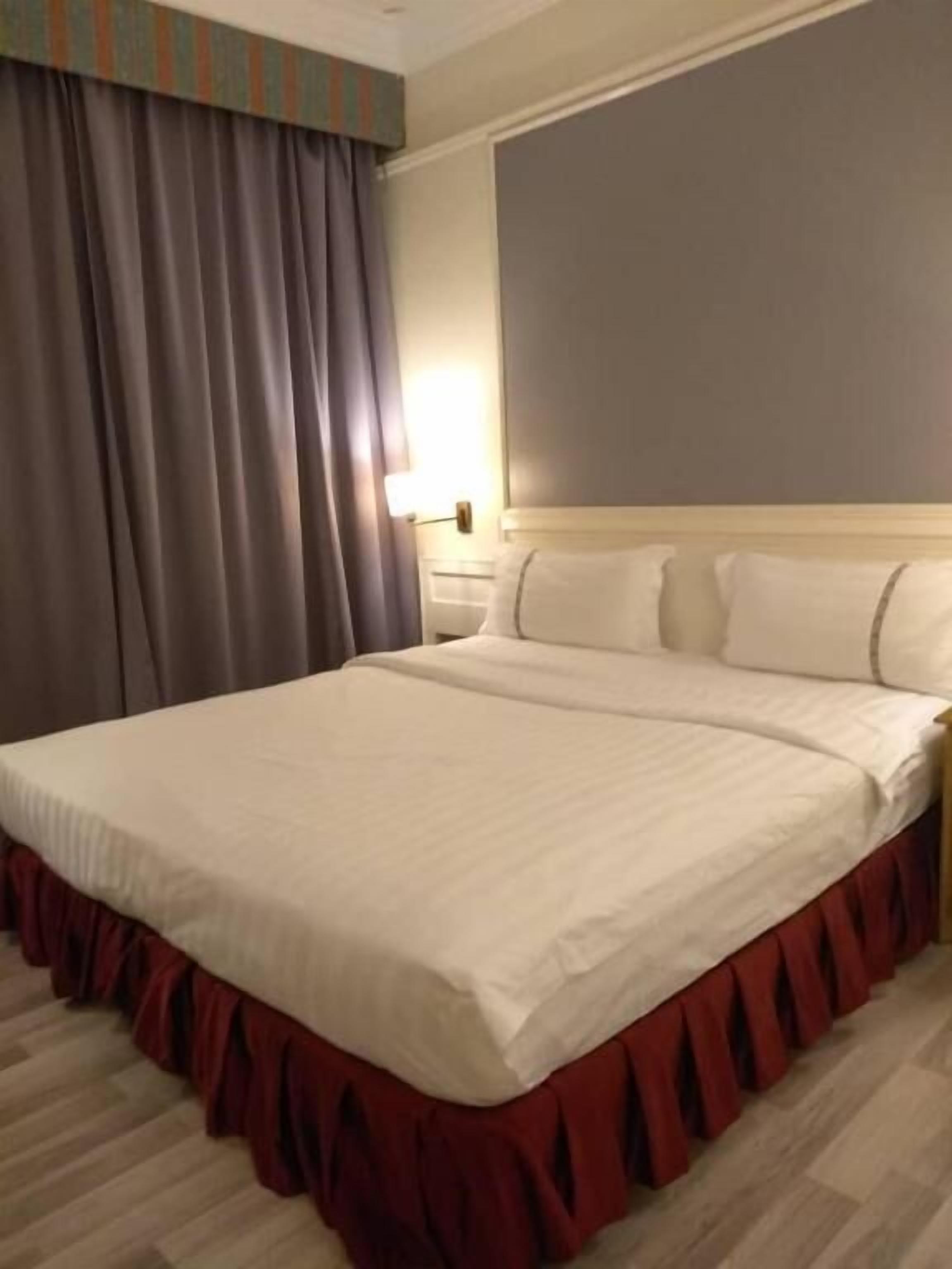 A neatly made bed with a white comforter and red skirt, a wooden headboard, and a nightstand with a lamp.