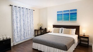 Une chambre à coucher comprenant un lit, deux tables de chevet, une commode, une fenêtre avec un rideau à motifs floraux et un mur orné de trois tableaux.