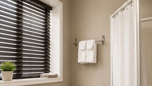 Une salle de bain comprenant des toilettes, un lavabo, une fenêtre avec des stores, un rideau de douche et une poubelle.