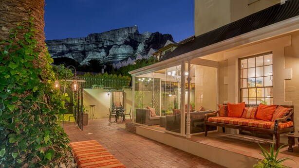 A patio area with a sofa and cushions, a table, and chairs. There is a glass enclosure with a view of a mountain. - Rosedene Guest House (Cape Town)
