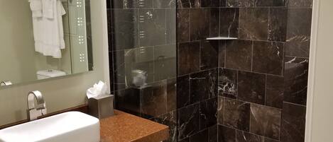 Un baño moderno con lavamanos blanco, cabina de ducha con mampara de vidrio y baldosas de mármol oscuro.