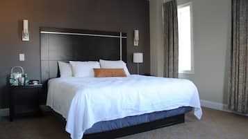 A hotel room with a large bed, dark headboard, two bedside tables, a nightstand, a window with curtains, and a patterned carpet.