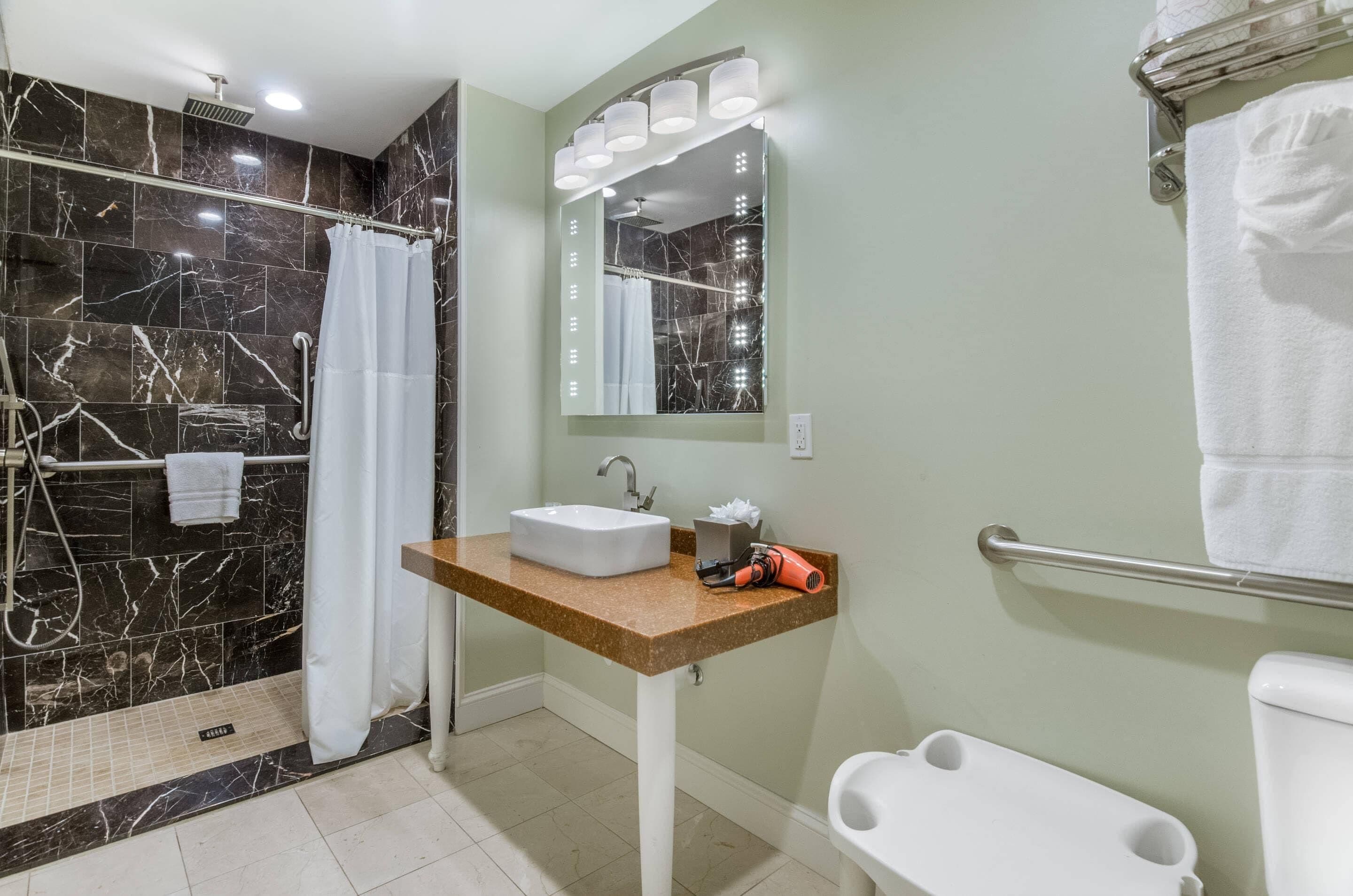 A bathroom with a marble shower, a sink with a towel rack, a mirror with lights, and a toilet.