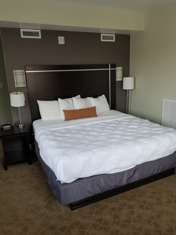 A hotel room with a large bed, two bedside tables, a nightstand, a lamp, and a wall-mounted air vent.