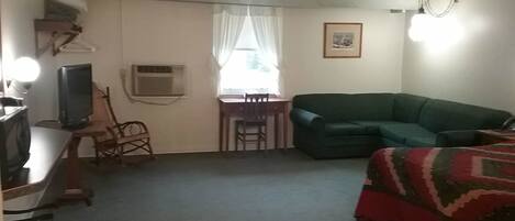 A room with a green sofa, a bed with a patterned blanket, a desk with a computer and a chair, an air conditioning unit, and a window with curtains.