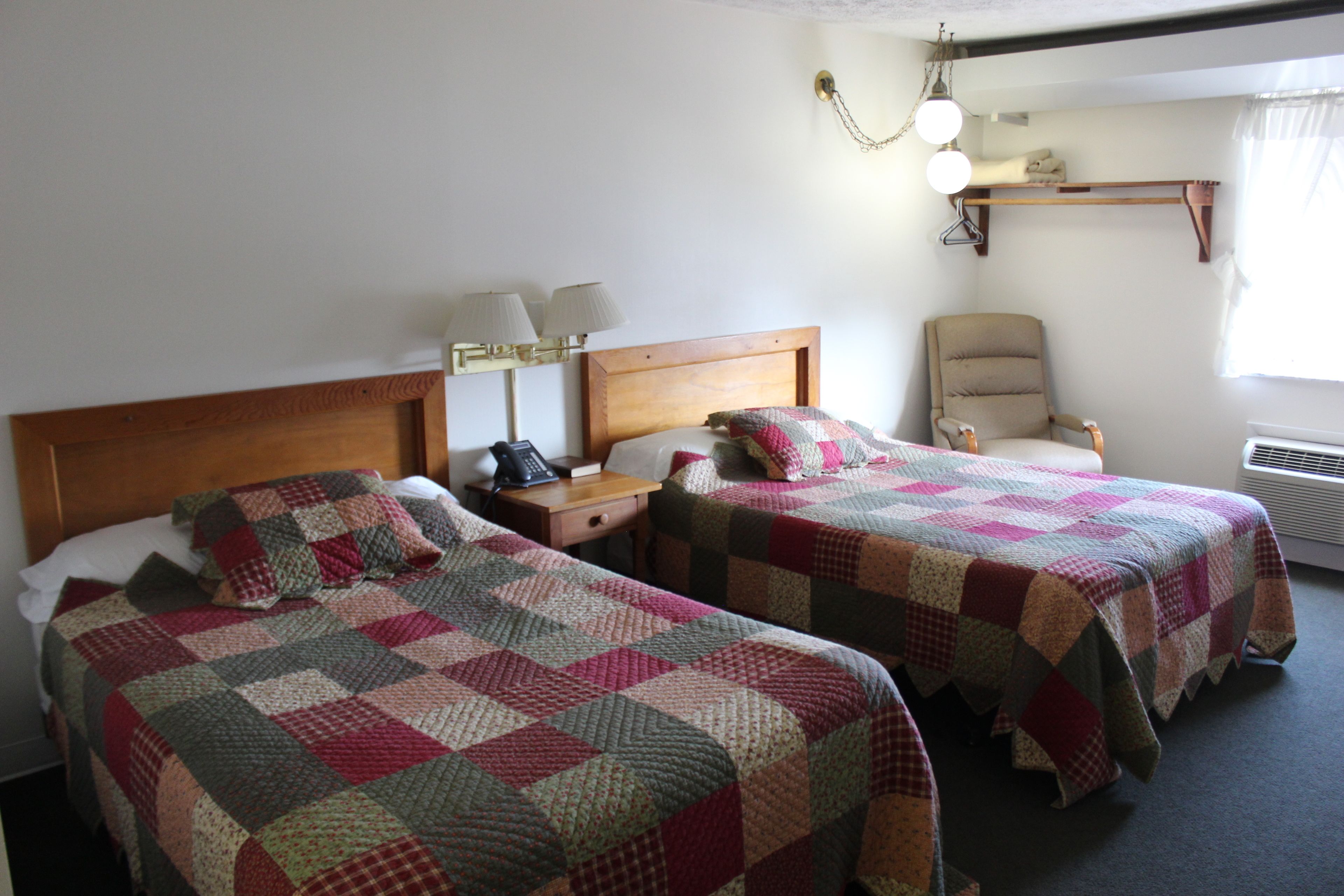 Una habitación con dos camas, cada una con colcha acolchada, una mesita de noche con lámpara, una silla y un estante sobre la cama.