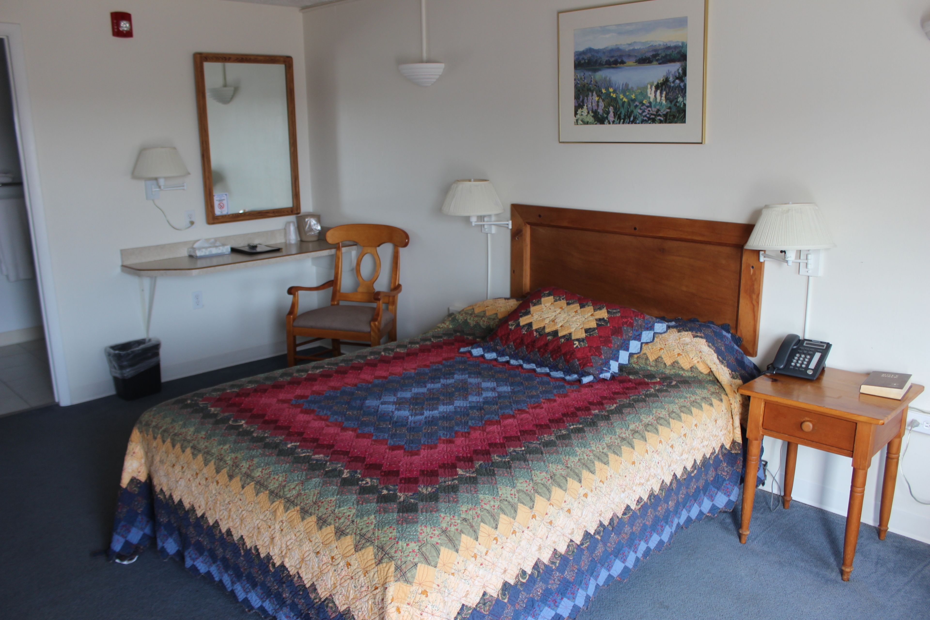 A neatly made bed with a colorful quilt, a wooden nightstand with a lamp, a chair, and a desk with a telephone and book.