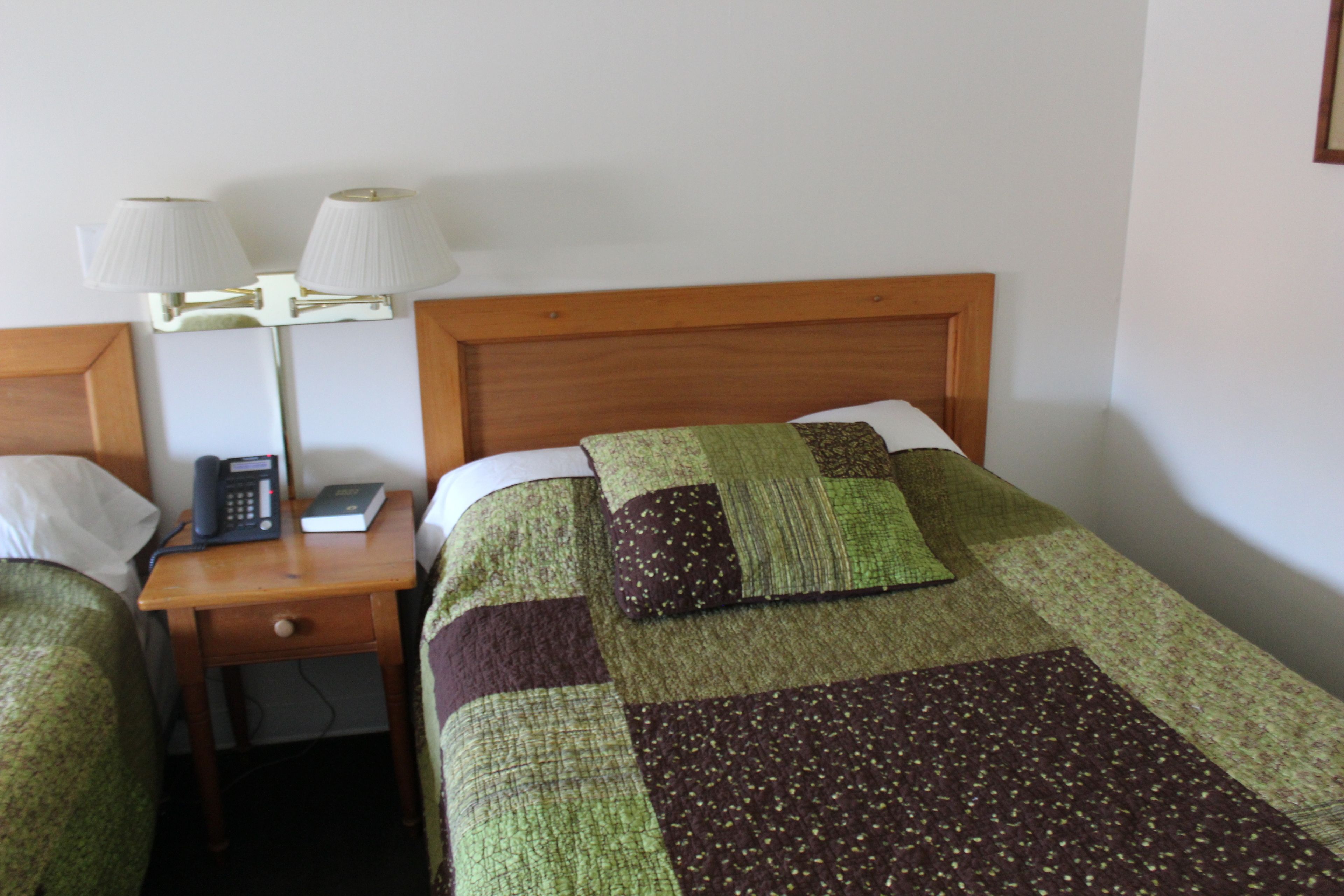 A hotel room with a wooden headboard, a bedside table with a lamp, a telephone, and a quilted bedspread.