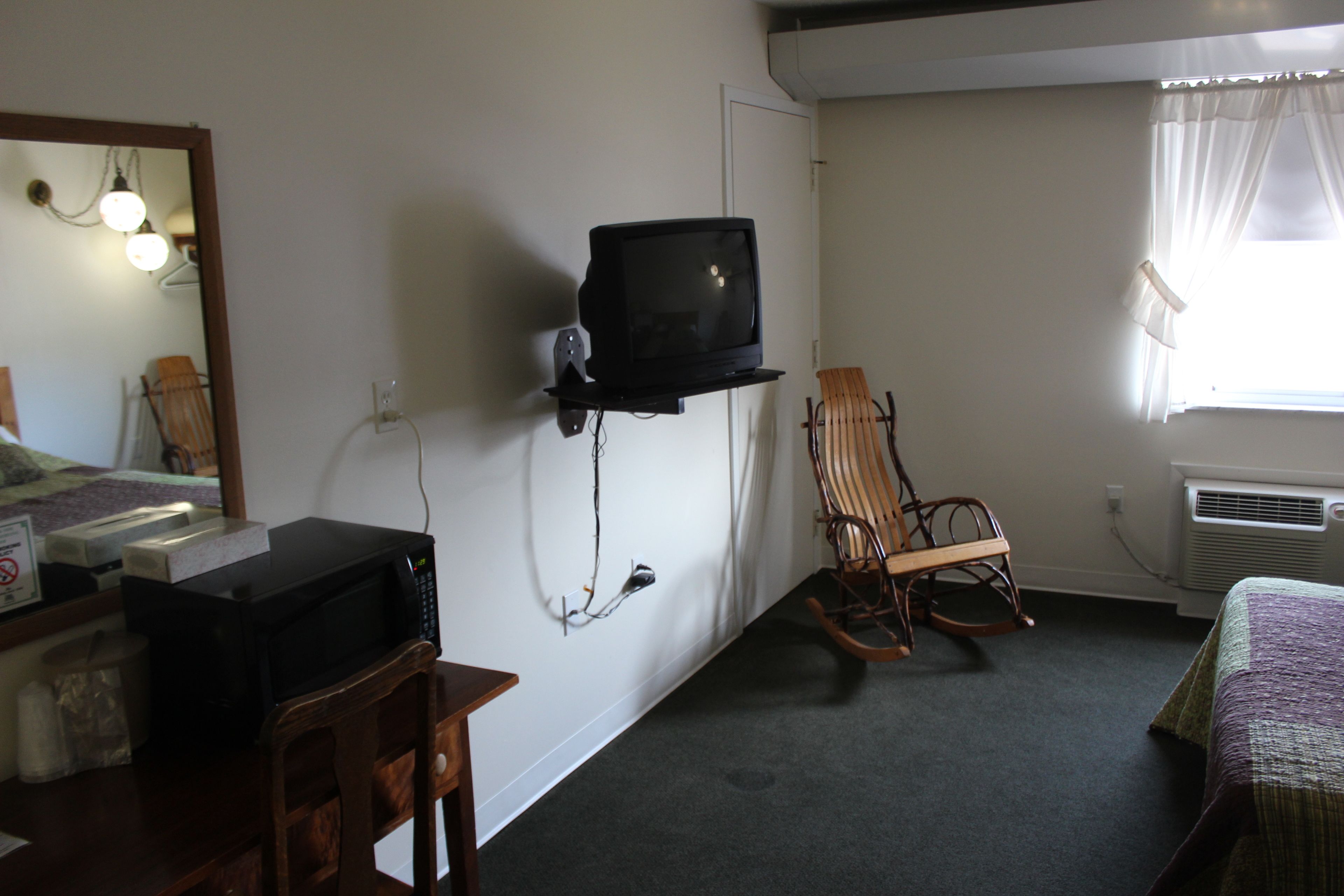 Una habitación con televisión montada en la pared, un sillón mecedora, una cama con colcha y un escritorio con microondas.
