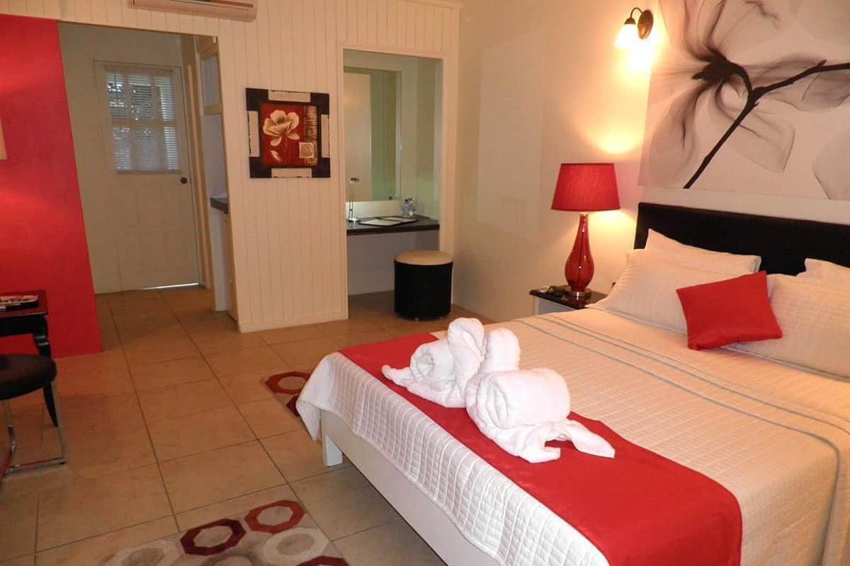 A hotel room with a bed, a red and white bedspread, a red lamp, a painting, and a bathroom visible through an open door.