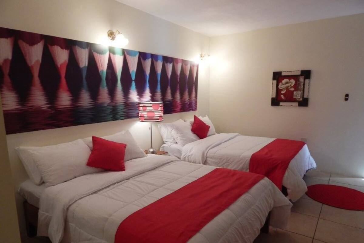 A hotel room with two beds, each with white bedding and red accents, a large abstract painting above the beds, and a small red rug on the floor.