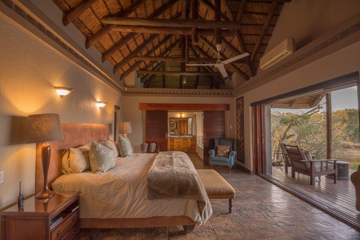 A spacious bedroom with a large bed, a wooden ceiling, and a view of a kitchen and outdoor area.