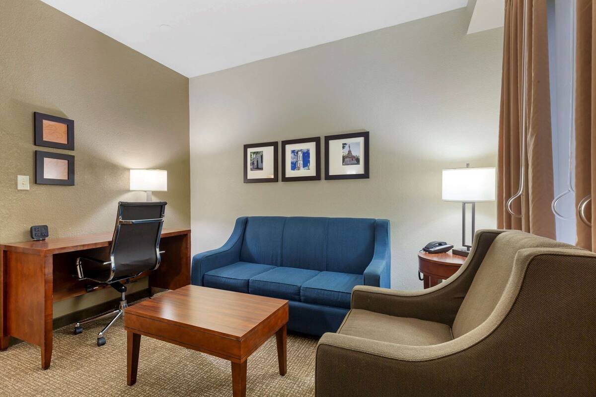 Una habitación de hotel con un escritorio de madera, un sofá azul, un sillón marrón y una mesita. Hay tres cuadros en la pared y una lámpara en el escritorio.