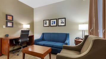 A hotel room with a wooden desk, a blue sofa, a brown armchair, and a small table. There are three framed pictures on the wall and a lamp on the desk.