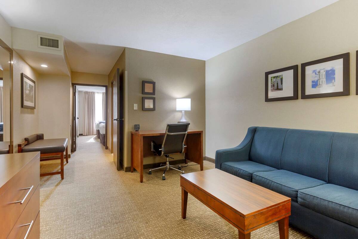 Habitación de hotel con un escritorio de madera, un sofá azul, una mesa de madera y un pasillo que conduce a otra habitación.