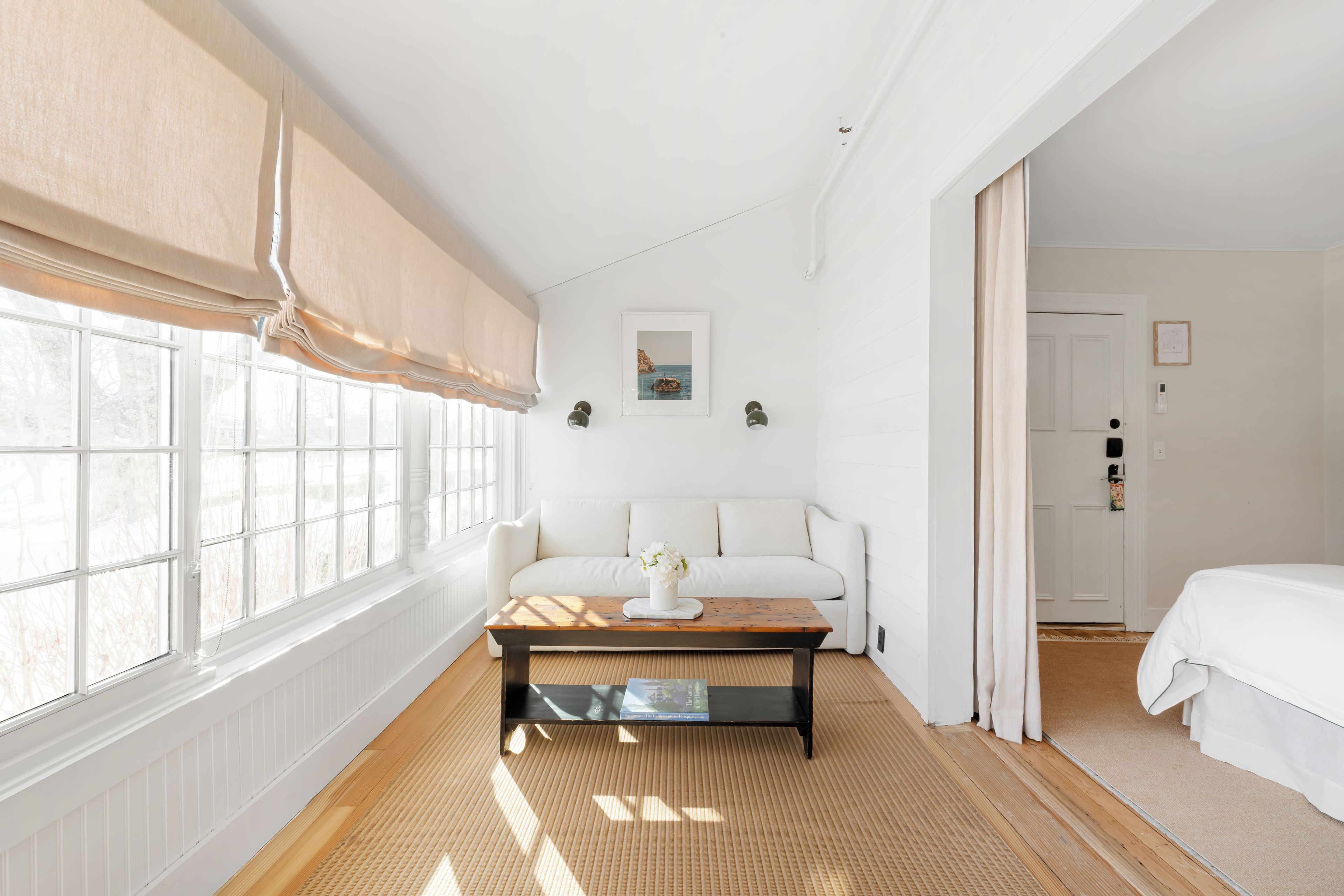 Une pièce avec un canapé blanc, une table basse en bois et une grande fenêtre avec des stores.