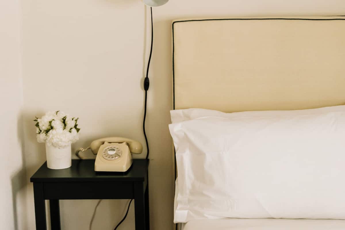 A neatly made bed with white linens, a small bedside table with a vintage telephone and a vase of flowers, and a wall-mounted lamp.