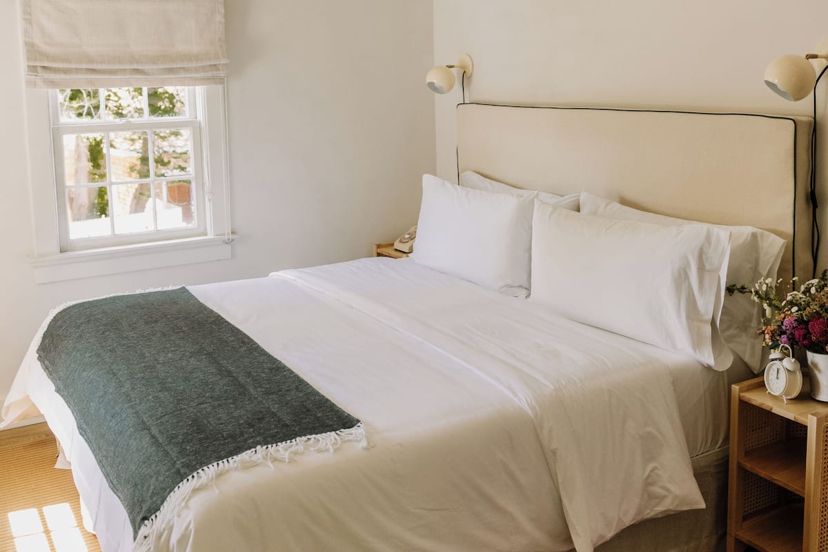 A neatly made bed with white linens, a wooden nightstand with a lamp, and a window with a view of greenery.
