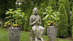 Une statue de jardin représentant une figure assise, avec deux plantes en pot de chaque côté.