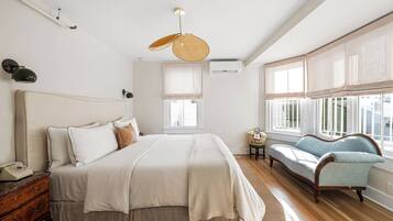 A bedroom with a large bed, bedside tables, a lamp, a hanging light fixture, an air conditioning unit, a window with curtains, and a sofa.