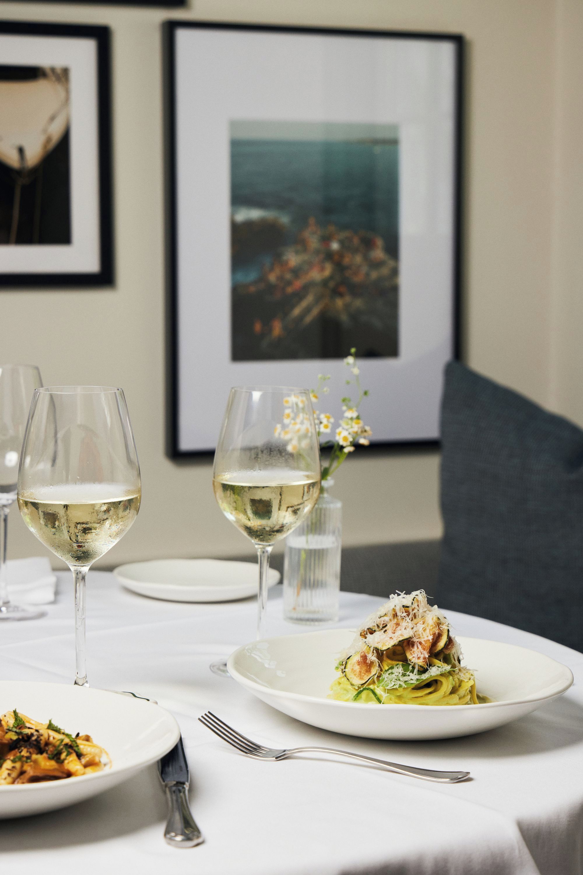 Une table à manger avec des assiettes remplies de nourriture, des verres de vin blanc et un tableau encadré accroché au mur.