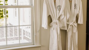 Une salle de bain avec deux peignoirs blancs suspendus à un porte-serviettes en bois, une fenêtre avec un store blanc et un autre porte-serviettes en bois sur lequel sont posées des serviettes.