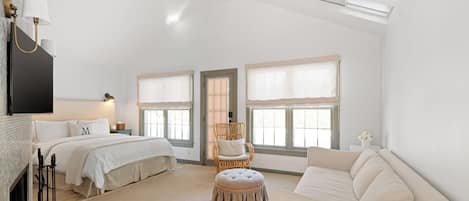A spacious bedroom with a large bed, a sitting area with a sofa, a chair, and a small ottoman. There are two windows with curtains, a fireplace, and a flat-screen TV mounted on the wall.