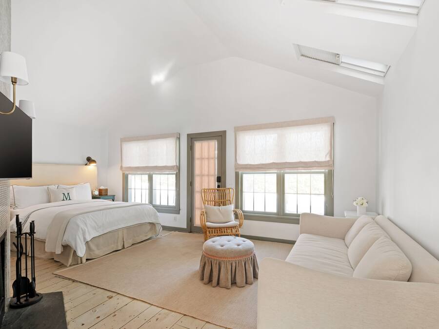Un dormitorio amplio con una cama grande, una zona de estar con un sofá, una silla y un pequeño reposapiés. Hay dos ventanas con cortinas, una chimenea y un televisor de pantalla plana montado en la pared.