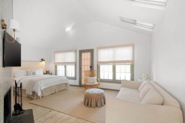 A spacious bedroom with a large bed, a sitting area with a sofa, a chair, and a small ottoman. There are two windows with curtains, a fireplace, and a flat-screen TV mounted on the wall.