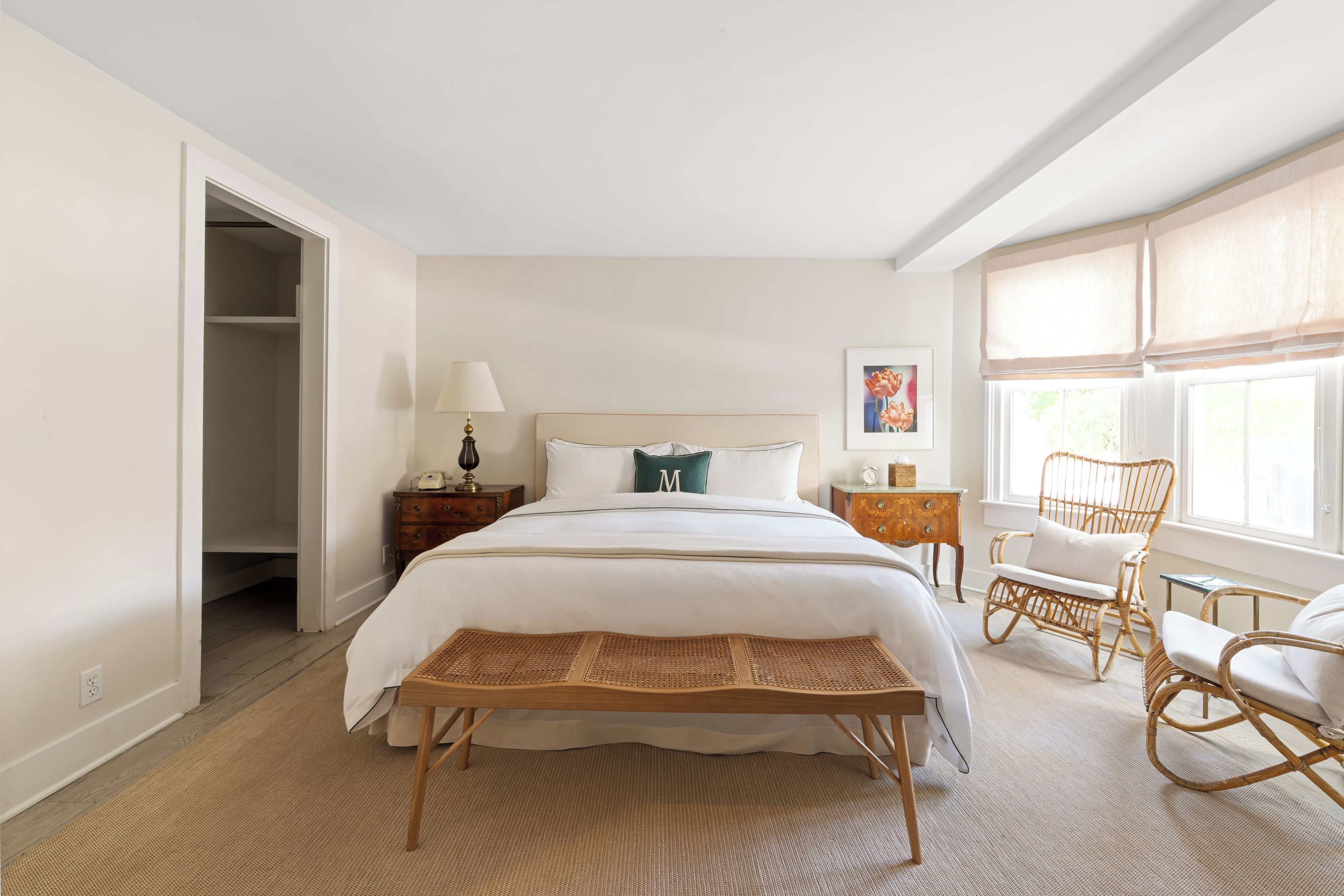 Een slaapkamer met een groot bed, een nachtkastje met een lamp, een rieten stoel en een raam met gordijnen.