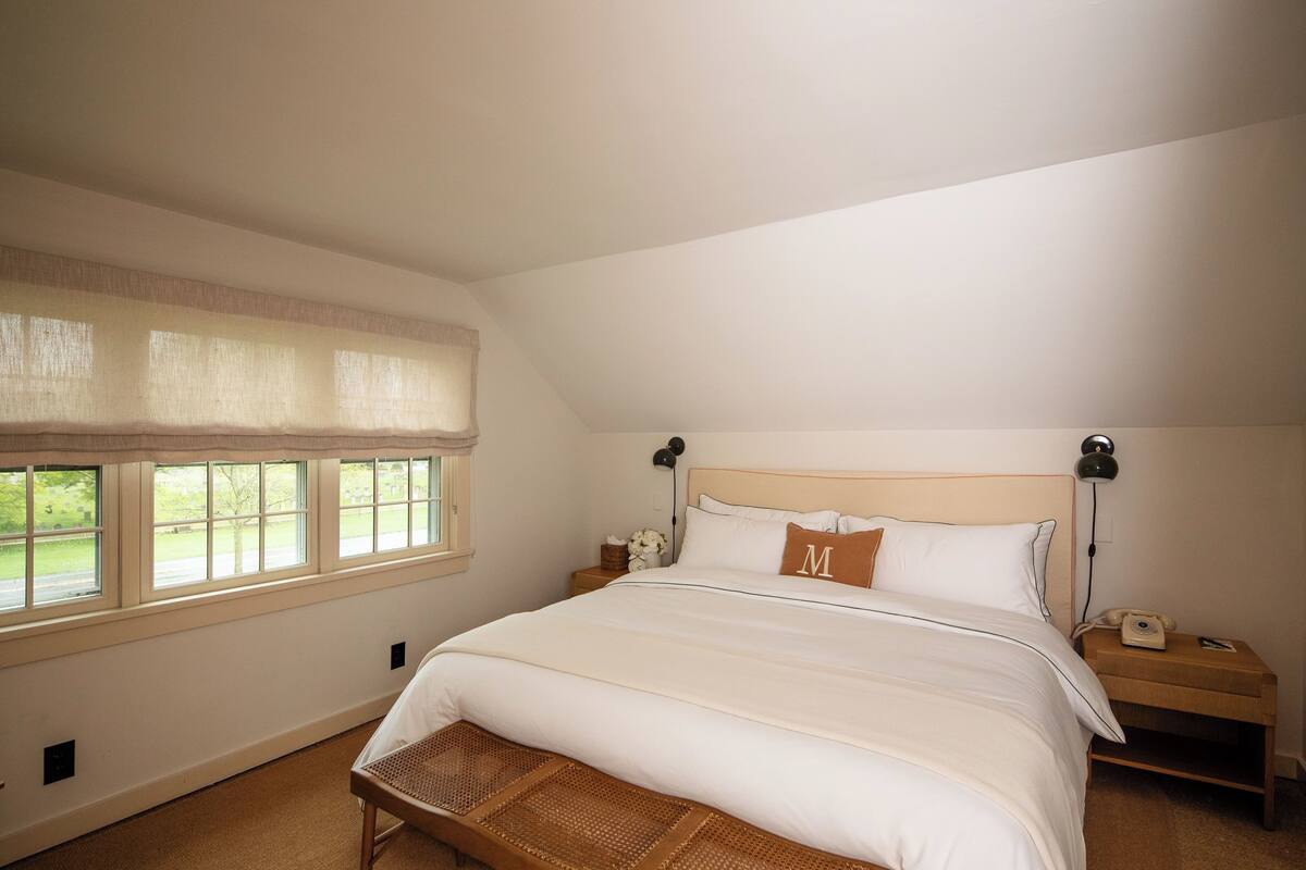 A bedroom with a large bed, a nightstand with a lamp, a wicker bench, and a window with a view of greenery.