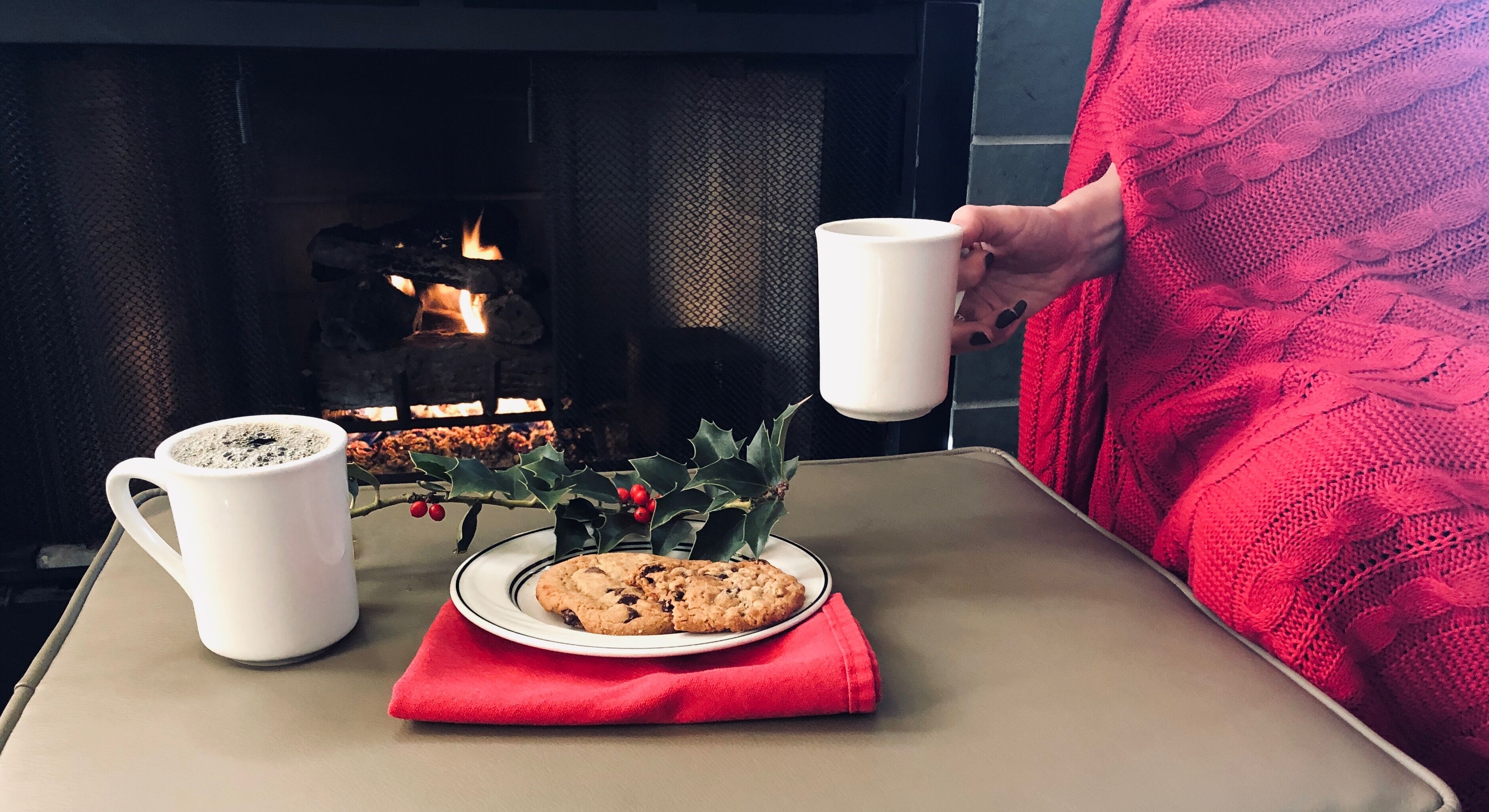 A table with a plate of cookies and two white mugs, a red napkin, and a decorative holly branch with berries.