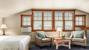 A hotel room with a bed, two armchairs, a small table with a vase of flowers, and large windows with blinds.
