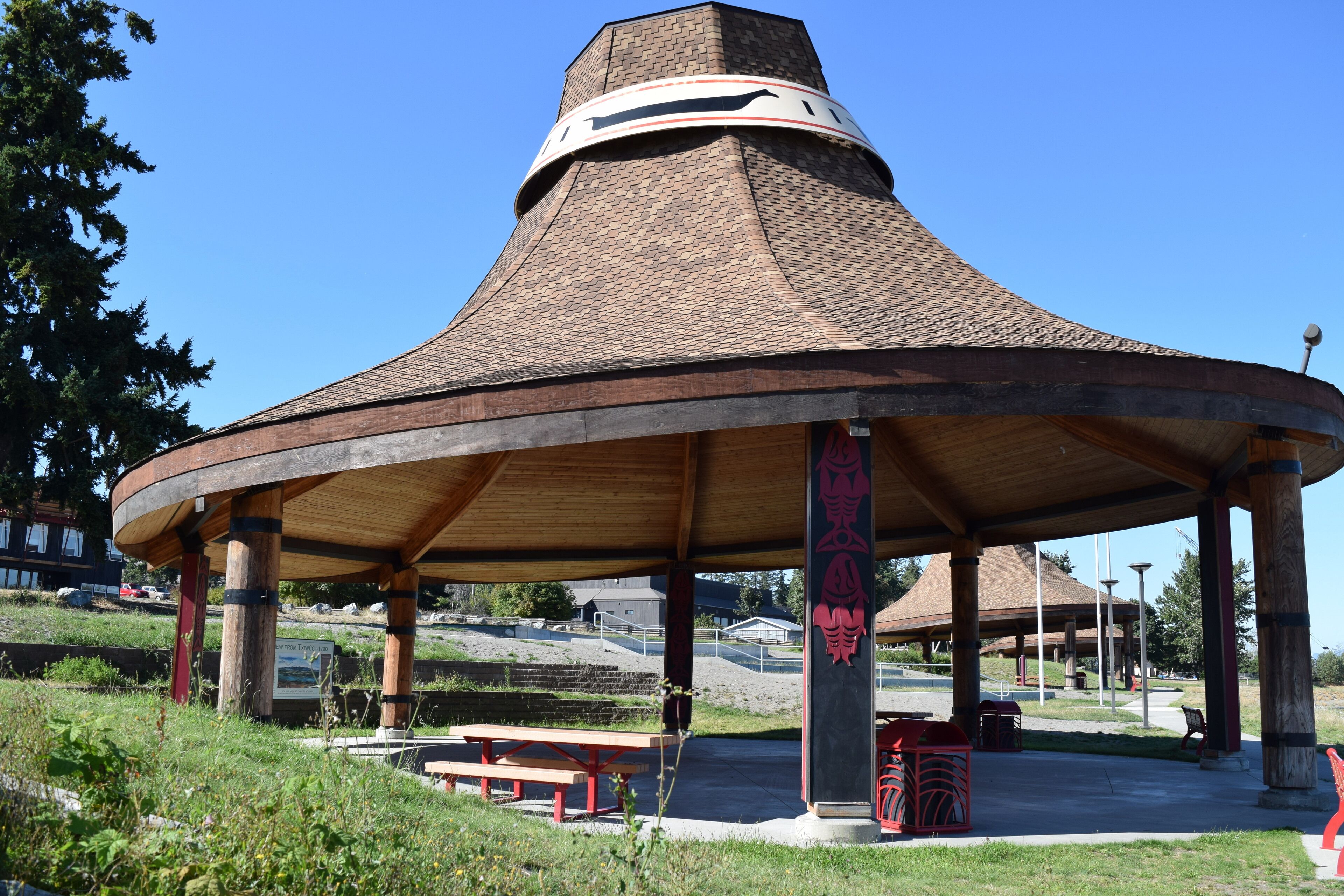 A pavilion with a conical roof, supported by wooden pillars, in a park setting with benches and picnic tables.