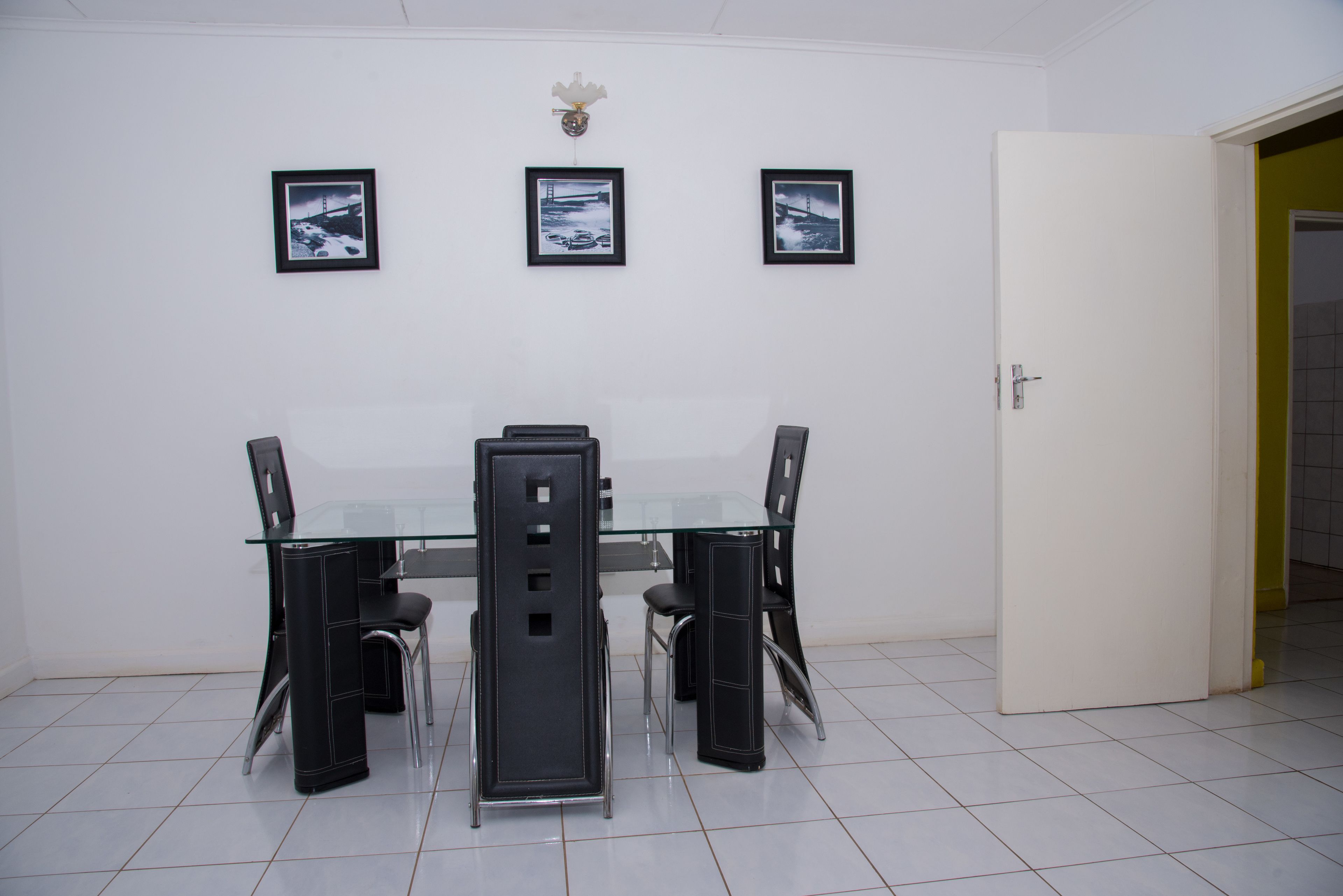 A dining area with a glass table and black chairs, three framed pictures on the wall, and a white door.