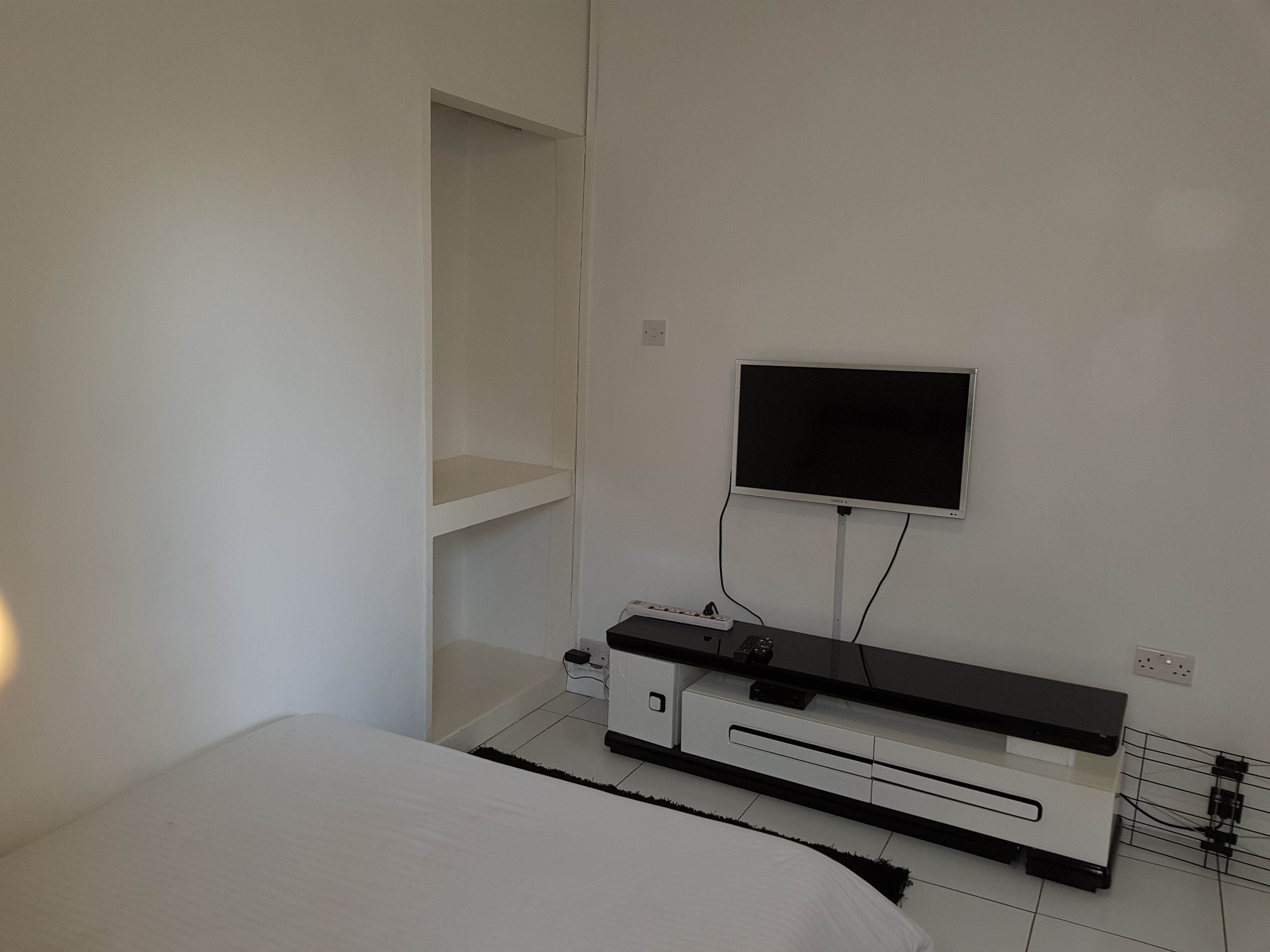 A bedroom with a bed, a TV mounted on the wall, a white wardrobe, and a bedside table.