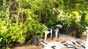 Un patio con suelo de baldosas, rodeado de abundante vegetación y una silla de plástico blanca.