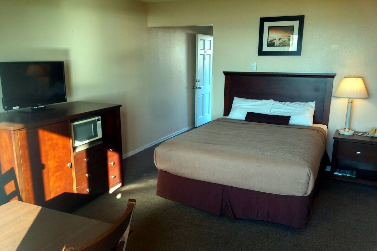 Quarto de hotel com uma cama, uma escrivaninha com televisão, uma luminária e uma porta que dá para outro cômodo.